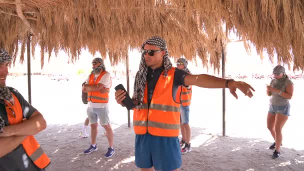 Egito, Hurgada, 09.08.2019: Feliz grupo sorridente de turista em coletes laranja, óculos escuros e headbands em pé sob capônia ouvindo música de um alto-falante sem fio — Vídeo de Stock