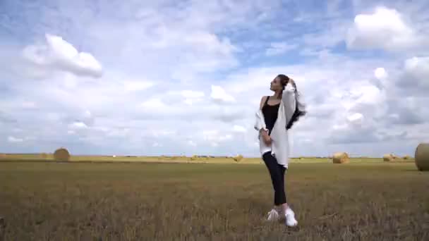 穿着黑色休闲装的美丽的黑发姑娘站在田野中间，她的头发在风中飘扬，蓝蓝的天空中衬托着白云 — 图库视频影像
