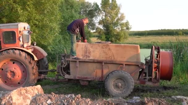 Muž v neformálním oblečení a masce stojí na traktoru u jezera a plní sud vodou — Stock video