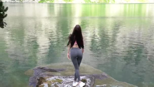 Jovem bela menina morena esportiva com cabelo solto vestido de roupas esportivas fica na grande pedra perto do incrível lago entre as montanhas — Vídeo de Stock