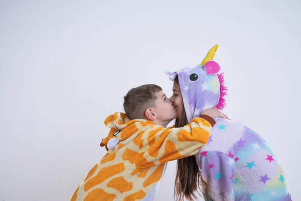 Brunett flicka i enhörning pyjamas står upphetsad medan hennes bror i giraffa pyjamas kramas och kyssa henne, isolerade över vit bakgrund — Stockfoto