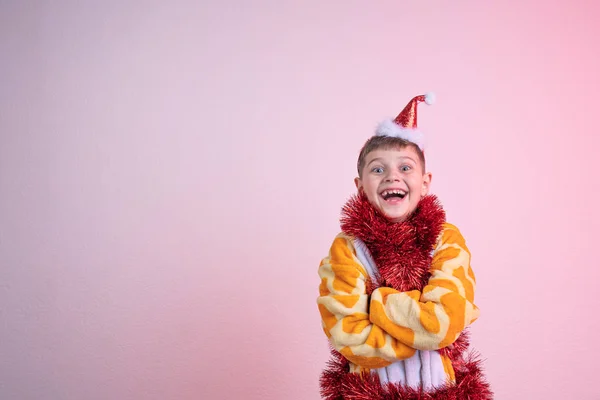 Excited happy laughing child, boy in red santa hat dressed in giraffe pajamas and wrapped in red new years rain, stands with cossed hands and looking at the camera isolated over pink background