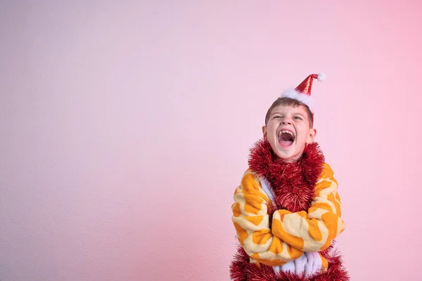 Spännande glad skrattar barn, pojke i röd tomte hatt klädd i giraff pyjamas och insvept i röda nya år regn, står med mysiga händer och öppnade munnen isolerad över rosa bakgrund, copyspace för — Stockfoto