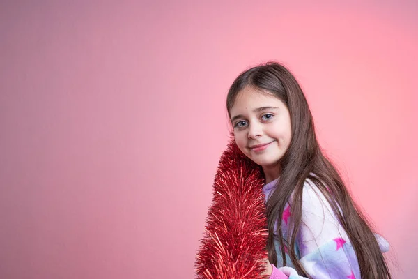 Hermosa chica morena con ojos azules y cabello suelto con lluvia de años nuevos rojos en el hombro, aislada sobre fondo rosa, mirando a la cámara, concepto de Navidad y Año Nuevo —  Fotos de Stock