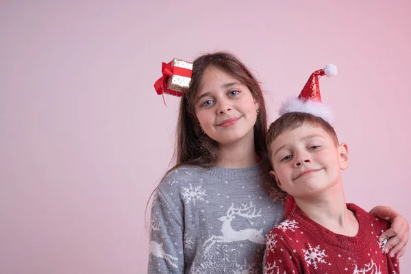 Primer plano retrato de hermosos niños en suéteres de santa y sombreros aislados sobre fondo rosa, hermana abrazando a su hermano, Feliz Año Nuevo y Feliz Navidad concepto —  Fotos de Stock