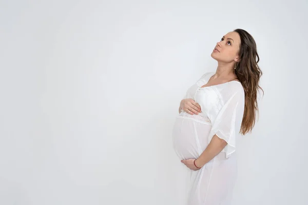 Portrait latéral de la jolie bunette fille enceinte aux cheveux lâches, vêtue de sous-vêtements blancs et tenant ses mains sur son ventre gonflé, isolée sur fond blanc, copyspace pour votre texte — Photo