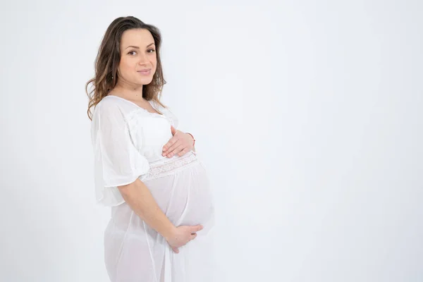 Portrait latéral de la jolie bunette fille enceinte aux cheveux lâches, vêtue de sous-vêtements blancs et tenant ses mains sur son ventre gonflé, isolée sur fond blanc, copyspace pour votre texte — Photo