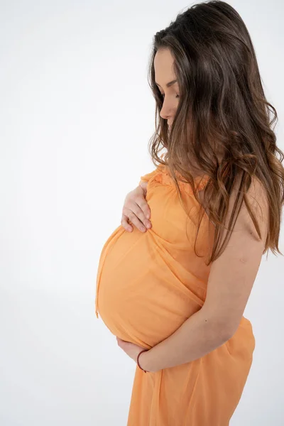 Morena jovem grávida com cabelo solto, vestindo vestido laranja e segura as mãos em sua barriga inchada, isolado sobre fundo branco — Fotografia de Stock