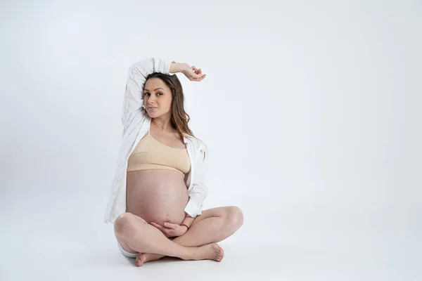 Belle brune aux cheveux lâches vêtue d'une chemise blanche assise par terre et tenant une main sur sa tête l'autre sur son ventre, isolée sur fond blanc, copyspace — Photo