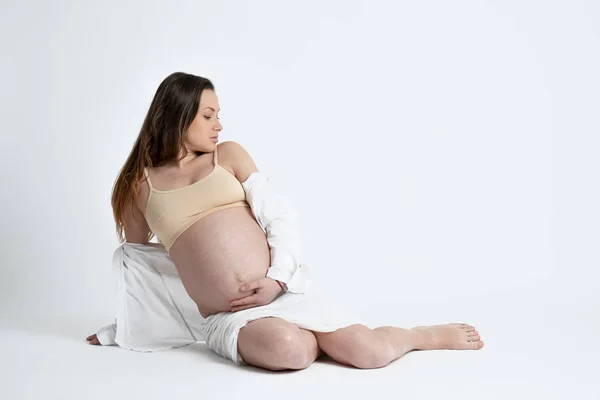 Menina morena grávida atraente em sutiã bege sentado no chão, inclinado na mão e olhando para o lado direito, espaço de cópia para o seu texto — Fotografia de Stock