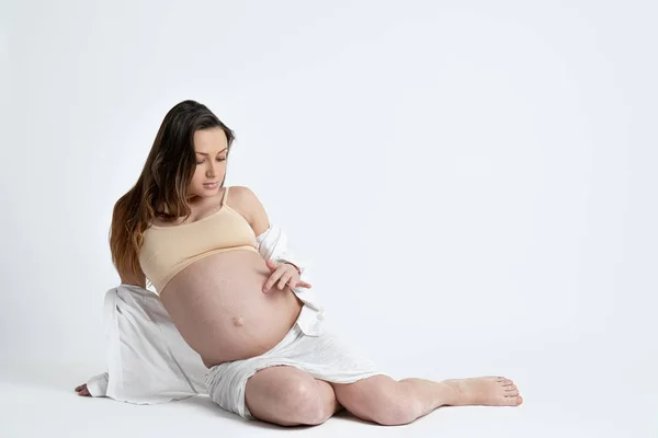 Estúdio foto de mulher bunette grávida em sutiã bege mentiras e olhando para sua barriga ao tocá-lo, copyspace para o seu texto — Fotografia de Stock
