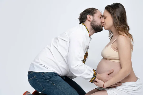 Young couple sitting on the ground, bearded man in white national shirt kissing his beautiful pregnant brunette wife, studio portrait — Stock Photo, Image