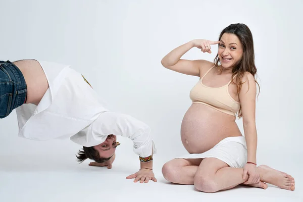 Sorrindo morena grávida mulher em sutiã bege sentado no chão, olhando para a câmera e torce um dedo perto da cabeça, enquanto seu marido, vestido com camisa branca, fica na posição de ponte — Fotografia de Stock