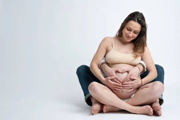 Jeune homme tenant ventre de sa femme enceinte faisant cœur. Femme enceinte et mari aimant embrassant le ventre à la maison. Coeur des mains par beau couple sur le ventre enceinte. Espace de copie, isolé — Photo