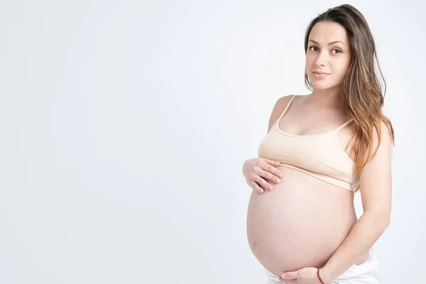 Attrayant jeune femme enceinte en soutien-gorge beige tient ses mains sur son ventre gonflé et en regardant la caméra, isolé sur fond blanc — Photo