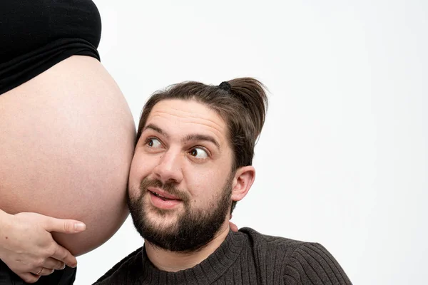 Bonito homem feliz em roupas casuais está ouvindo suas esposas grávidas barriga, isolado sobre fundo branco, copyspace para o seu texto — Fotografia de Stock