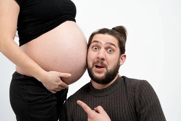 Joyful, playful happy bearded man in casual clothes is listening with funny face to his pregnant wifes tummy, isolated over white background, copyspace for your text — Stock Photo, Image