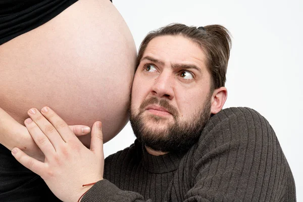 Embarrassé homme barbu en vêtements décontractés écoute ses femmes enceintes ventre, isolé sur fond blanc — Photo