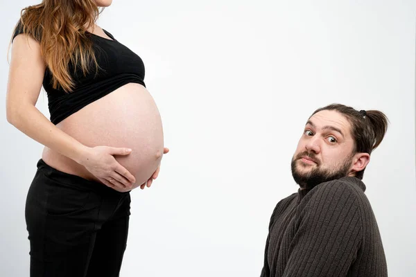 Embarrassed young bearded man looking at the camera while his pregnant whife in black clothes stands near him and holds her hands on her belly, isolated over white background — Stock Photo, Image