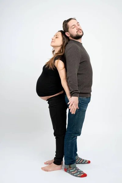 Молода мила пара в повсякденному одязі, бородатий чоловік і вагітна жінка, стоячи спиною до спини з закритими очима і тримаючи руки разом, ізольовані на білому тлі — стокове фото