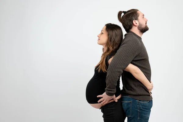 Мила сім'я, молода щаслива пара в повсякденному одязі, бородатий чоловік і його вагітна дружина стоять спиною і тримають руки один на одному живіт, копія — стокове фото