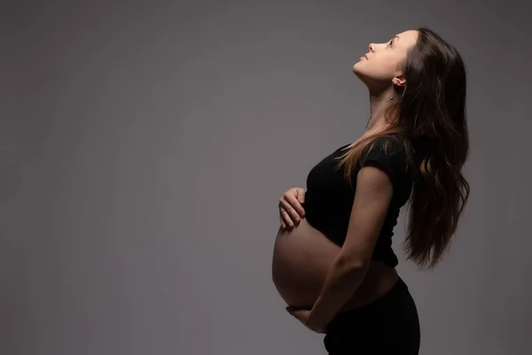 Side view portrait of the beautiful pregnant woman with loose hair that looking up and holds hands on her swollen belly, isolated over dark background, copyspace for your text