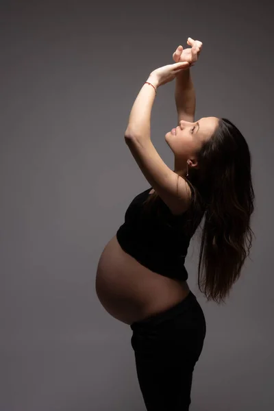 Visão lateral retrato da bela mulher grávida com cabelos soltos que olhando para cima e segura as mãos levantadas, isolado sobre fundo escuro — Fotografia de Stock