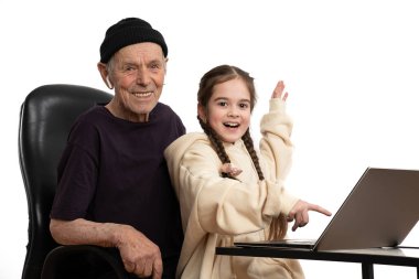 Diz üstü bilgisayarla bej kapüşon giyen mutlu torun siyah şapkalı ve tişörtlü şık dedesiyle birlikte beyaz arka planda izole edilmiş bir şekilde vakit geçiriyor.