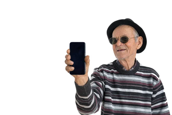 Gelukkige stijlvolle tandeloze oude man met zonnebril en zwarte hoed houdt telefoon in zijn hand en kijkt ernaar, geïsoleerd over witte achtergrond, copyspace voor uw tekst — Stockfoto