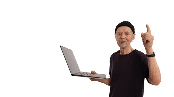 Feliz anciano maduro con sombrero negro y camiseta apuntando hacia arriba y mirando a la cámara, sostiene la computadora portátil en su mano y sonríe aislado sobre el fondo blanco — Foto de Stock