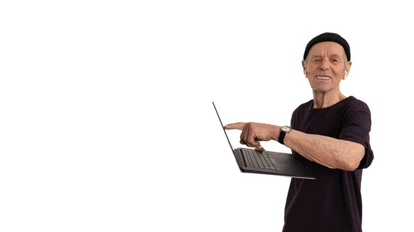 Happy old man pencioner in black hat and t-shirt pointing at blank screen of laptop computer and looking at the camera, holds laptop computer in his hand and smiling isolated over white background — Stock Photo, Image