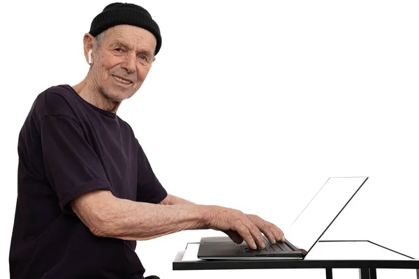 Happy old man in black hat, t-shirt and white wireless headphones, senior programmer typing on the keyboard and looking at the camera, works at a laptop, isolated over white background — Stock Photo, Image