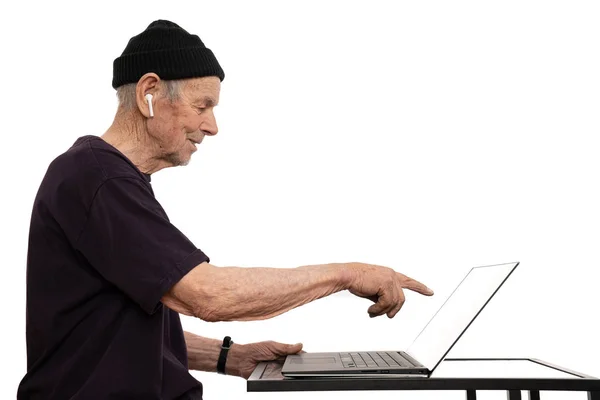 Discomposed old man in black hat, t-shirt and white wireless headphones, senior chat administrator pointing at blank screen of laptop computer, isolated over white background — Stock Photo, Image