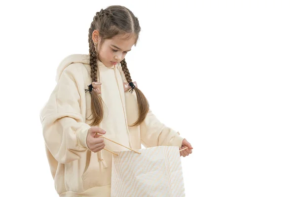Sorglig, olycklig liten flicka med flätor klädda i beige hoodie håller shoping paket i händerna och tittar inuti, isolerade över vit bakgrund — Stockfoto