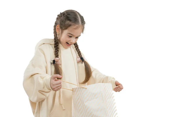 Glad, upphetsad liten flicka med flätor klädda i beige hoodie håller shoping paket i händerna och tittar inuti, isolerade över vit bakgrund — Stockfoto