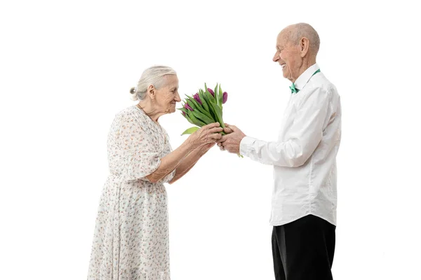 Velha mulher de vestido branco cheirando flores que seu marido sênior segura em suas mãos dá-lhe buquê enquanto celebra o dia das mulheres, isolado sobre fundo branco — Fotografia de Stock