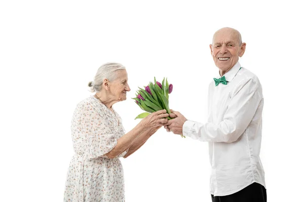 Velha mulher de vestido branco cheirando flores que seu marido sênior segura em suas mãos dá-lhe buquê enquanto celebra o dia das mulheres, isolado sobre fundo branco — Fotografia de Stock