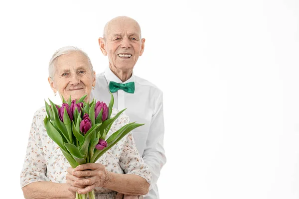 Derékig érő portré a boldog, mosolygó idős párról, aki fehér ingben ölelgeti a feleségét fehér ruhában, aki virágokat tart a kezében, mindketten a fehér háttér felett elszigetelt kamerát nézik. — Stock Fotó