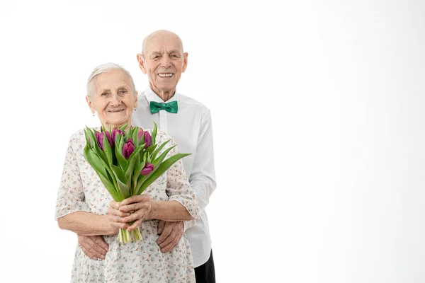 Retrato de la cintura hacia arriba de los abuelos sonrientes felices, senior en camisa blanca abrazando a su esposa en vestido blanco que sostiene las flores en las manos, ambos están mirando a la cámara aislada sobre fondo blanco — Foto de Stock