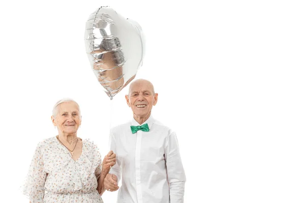 Duas pessoas despreocupadas, descuidadas, positivamente casal com cabelos grisalhos abraçar uns aos outros em interior branco brilhante e olhar para a câmera, segurando balão de prata, copyspace para o seu texto — Fotografia de Stock