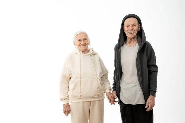 Feliz casal sênior elegante em roupas casuais com airpods brancos em seus ouvidos, homem velho e mulher de capuz de mãos dadas e olhando para a câmera, isolado sobre fundo branco — Fotografia de Stock