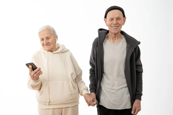 Alegre elegante sênior em capuz e chapéu preto segura a mão de sua esposa que olhando para o telefone, isolado sobre fundo branco — Fotografia de Stock