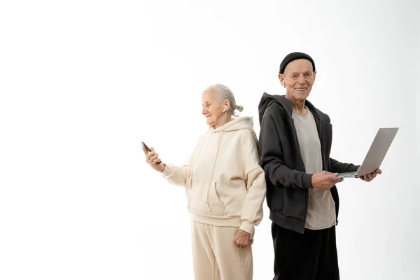 Pessoas idosas sorridentes felizes usando a tecnologia moderna, um computador portátil e um smartphone isolado no fundo branco, homem e mulher elegantes em roupas casuais, sênior em chapéu preto detém laptop e — Fotografia de Stock