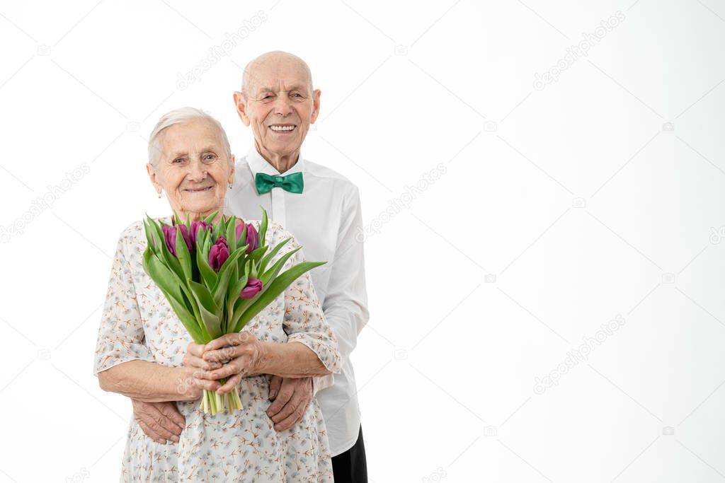 Waist up portrait of the happy smiling grandparents, senior in white shirt hugging his wife in white dress that holds flowers in hands, both are looking at the camera isolated over white background