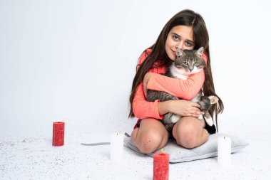 Küçük esmer kız yastığın üzerinde oturuyor, kediye sarılıyor ve kameraya bakıyor, kırmızı ve beyaz mumlar beyaz arka planda izole bir şekilde etrafa konuyor.
