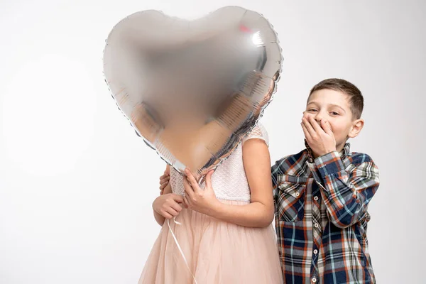 Attractive little couple, girl in pink dress hide her face behinde silver balloon and little boy in checkered shirt hide his face with his hand, isolated over grey background — Stockfoto