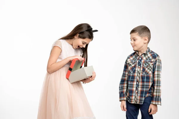 Happy little brunette girl in beige dress open box with christmas present her brother in shirt loooking at her, isolated over white background — Stockfoto