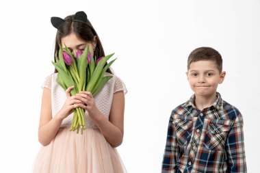Confident little boy in shirt looking at the camera, his attractive sister in beige dress hide her face with purple tulips, isolated over white background