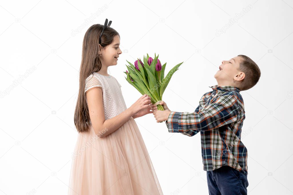 Little boy in a shirt giving a purple tullips to fashionable child girl in beige dress isolated on white background - celebrating womans day, St. valentines day, birthday