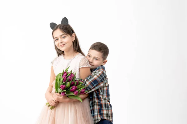 Schöner kleiner Junge im Hemd umarmt seine fröhliche süße Schwester im beigen Kleid, brünettes Mädchen hält lila Blumen in ihren Händen und schaut in die Kamera, isoliert vor weißem Hintergrund — Stockfoto